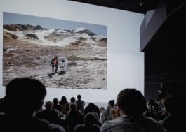 A photograph showing an analog astronaut sitting on a chair in alpine environment being projected within the Deep Space Theater at Ars Electronica Festival 2017 