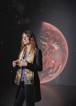 Astronomer Lisa Kaltenegger is standing in front of a projection of a Red-Tainted Expolanet. She has long, reddish Hair and is wearing a black blazer, blue jeans anda silk scarf reminiscent of a painting by Gustav Klimt.