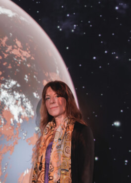 Astronomer Lisa Kaltenegger is standing in front of a projection of a Red-Tainted Expolanet. She has long, reddish Hair and is wearing a black blazer, blue jeans anda silk scarf reminiscent of a painting by Gustav Klimt.