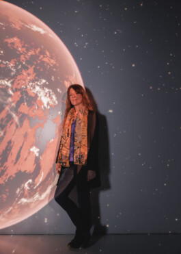 Astronomer Lisa Kaltenegger is standing in front of a projection of a Red-Tainted Expolanet. She has long, reddish Hair and is wearing a black blazer, blue jeans anda silk scarf reminiscent of a painting by Gustav Klimt.