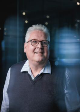 AI Researcher Sepp Hochreiter in a futuristic setting with lots of blue light streaks and reflections. He's wearing a black vest over a white shirt and blue jeans. He has white hair and is wearing glasses. 